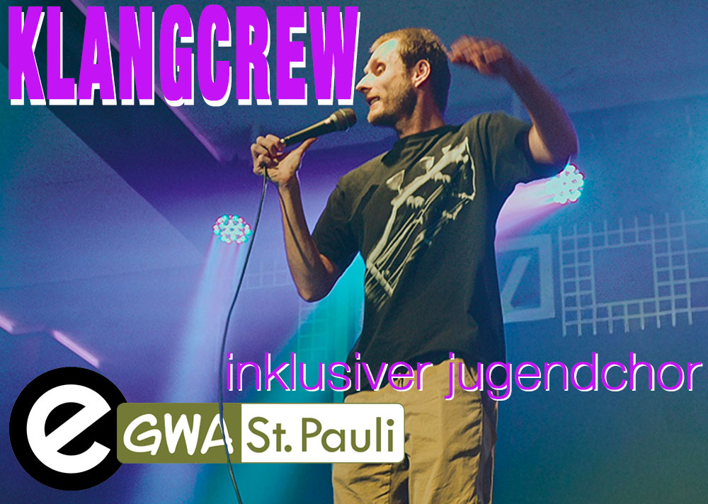 Der Chorleiter Nick steht auf einer Bühne mit Mikrofon in der Hand. Daneben das Logo von KlangCrew, der Esche und GWA St. Pauli