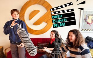Jugendliche von DREH DEINEN FILM! e.V. haben auf der Couch im Esche Jugendkunsthaus eine Filmklappe und Mikrofon in der Hand.