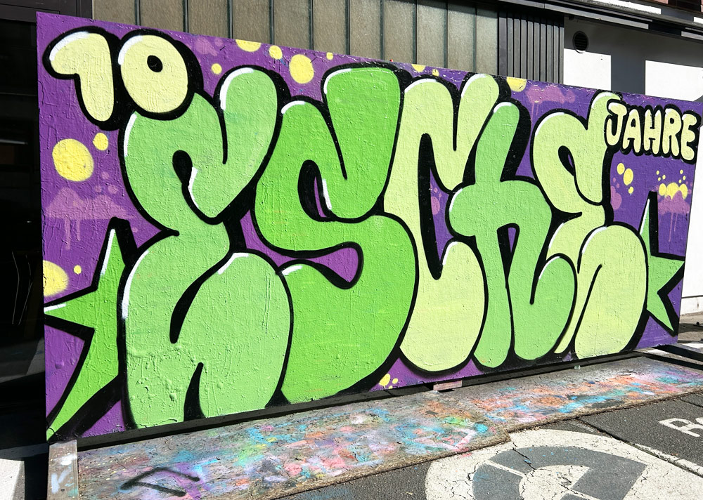 Ein großes 10-Jahre-Graffiti auf der Holzwand vor der Esche, davor stehen eine Hndvoll Jugendlicher.