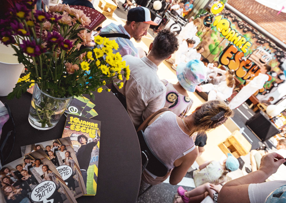 Publikum vor Jugendlichen bei unserem Sommerfest. Im Vordergrund steht eine Vase und Flyer der Esche liegen auf einem Stehtisch aus.